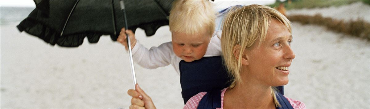 En mamma på stranden med ett barn på ryggen och paraplyt uppe.